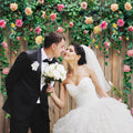 A bride and groom, dressed in formal wedding attire, kiss in front of a wooden fence adorned with Spring Floral Wood Plank Photoshoot Flowers Backdrop -ideasbackdrop and green vines. The bride holds a bouquet of white flowers, creating a stunning floral backdrop curated by expert event decorators from ideasbackdrop.