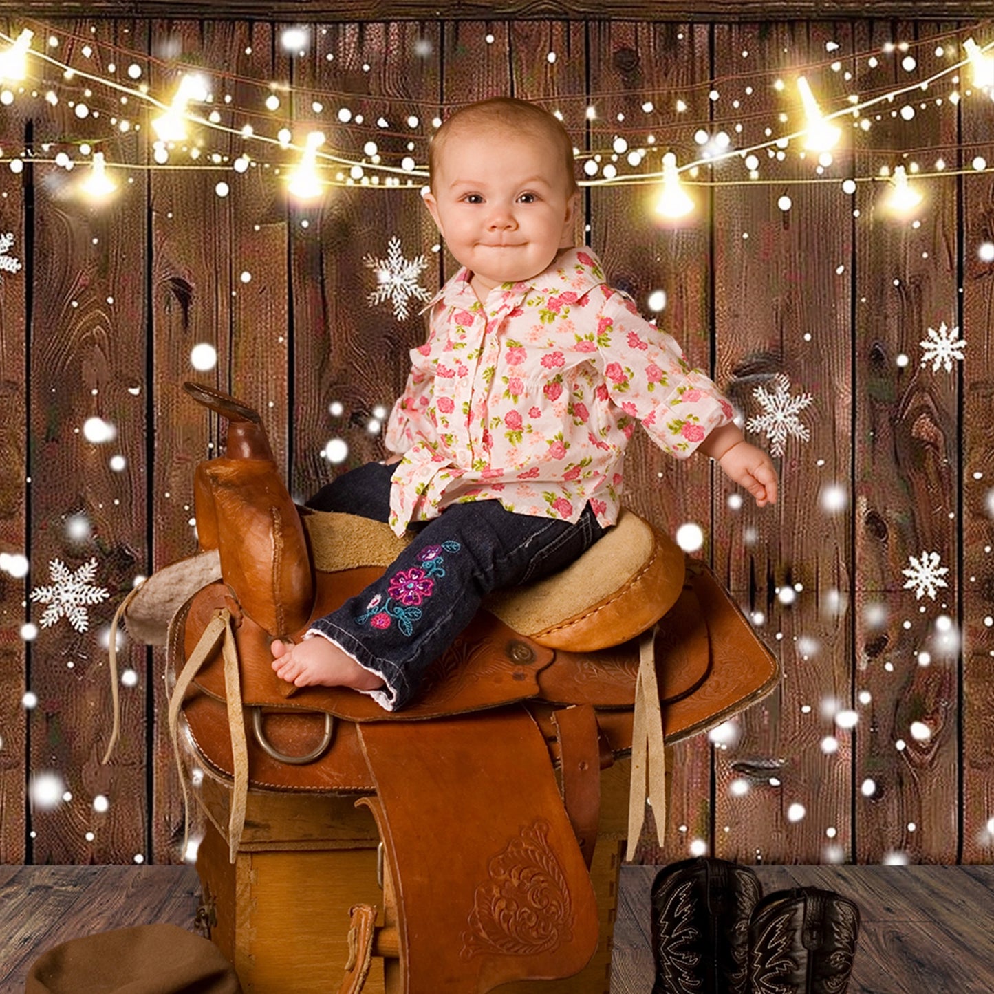 A baby sits on a saddle, wearing a floral shirt and jeans, in front of an ideasbackdrop Rustic Glitter Background Wood Backdrop. There are wintery string lights and snowflake decorations in the background. Cowboy boots are placed on the floor next to the saddle, adding to the vintage design charm of the scene.