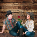 A young man and woman dressed in cowboy hats and casual clothing sit on the wooden floor against a Wooden Boards Photography Wood Backdrop-ideasbackdrop by ideasbackdrop. The woman holds a red rose while they smile at each other, evoking a nostalgic atmosphere.