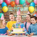 Five children wearing party hats smile and stand around a birthday cake with lit candles. Colorful balloons and streamers decorate the wooden background, which features a realistic wood grain pattern in an HD backdrop. The vibrant scene is set against the Colorful Carnival Balloons Wood Backdrop-ideasbackdrop by ideasbackdrop.