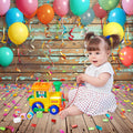 A toddler sits on a wooden floor, the realistic wood grain pattern adding charm as they play with a toy truck. Colorful balloons and confetti decorate the scene against an HD ideasbackdrop Colorful Carnival Balloons Wood Backdrop-ideasbackdrop.