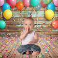 A baby in a white tank top and jeans sits on a wooden floor sprinkled with colorful confetti, surrounded by colorful balloons and streamers hanging from a Colorful Carnival Balloons Wood Backdrop-ideasbackdrop. The baby has one finger in their mouth.