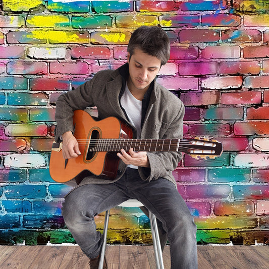 A person plays an acoustic guitar while sitting on a stool in front of a colorful, graffiti-covered brick wall backdrop that showcases stunning detail, thanks to high-resolution printing technology from the 7x5ft Fabric Graffiti Brick Wall Backdrop for Photography Hip Hop Disco Background Party Banner Portraits Photoshoot Props by ideasbackdrop.