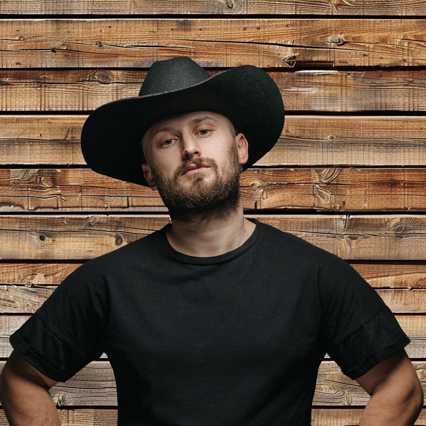 Man with a beard and mustache, dressed in a black t-shirt and black cowboy hat, stands in front of an ideasbackdrop 7x5ft Wood Backdrops for Photography Worn Wooden Boards Background Brown Photo Wall Photo Studio, perfect for high-resolution printing.