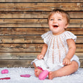 A baby wearing a white knitted outfit sits on a white rug in front of ideasbackdrop's 7x5ft Wood Backdrops for Photography Worn Wooden Boards Background Brown Photo Wall Photo Studio, smiling and holding a pink toy. Pink objects are scattered around.