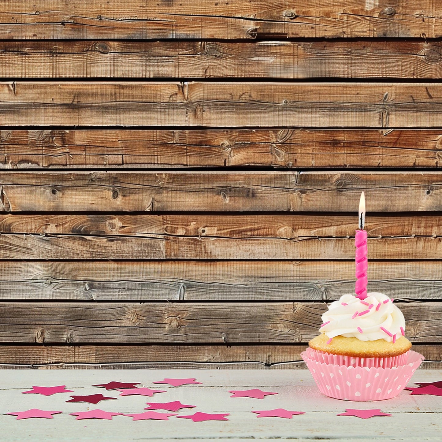 A single cupcake with white frosting and a pink candle is placed on a white surface. Pink star-shaped confetti is scattered around. The ideasbackdrop 7x5ft Wood Backdrops for Photography Worn Wooden Boards Background Brown Photo Wall Photo Studio enhances the charm of this delightful scene.