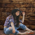 A person in a plaid shirt and jeans sits barefoot against a ideasbackdrop Rustic Wood Wall Backdrop Natural Brown Wooden Board Photography Background Baby Shower Birthday Party Cake Table Decor, wearing a cowboy hat and holding a small bunch of flowers.
