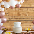 A white tiered cake on a wooden table with two plates of desserts on either side. A balloon garland in shades of white, gold, and rose gold decorates the 7x5ft Retro Wood Graduate Wall Background Wooden Backdrop Studio Props for Baby Shower Birthday Photography by ideasbackdrop.