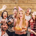 A group of people wearing party hats are celebrating with sparklers. A woman in the center holds a birthday cake. Everyone appears happy and festive against an ideasbackdrop 7x5ft Retro Wood Graduate Wall Background Wooden Backdrop Studio Props for Baby Shower Birthday Photography, which adds a warm, rustic charm to the scene.