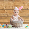 A baby wearing a pink bunny ears headband sits in a wicker basket. Colorful eggs and a stuffed toy are scattered around on a white furry surface, with an ideasbackdrop 7x5ft Retro Wood Graduate Wall Background Wooden Backdrop Studio Props for Baby Shower Birthday Photography that features high-resolution printing for added detail.