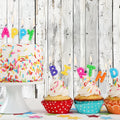 A birthday cake with sprinkles and candles spelling "HAPPY" next to three cupcakes with candles spelling "BIRTHDAY," topped with frosting and sprinkles, set against an ideasbackdrop Wood Backdrop Retro Rustic White Gray Wooden Floor Background for Photography Kids Photo Booth Video Shoot Studio Prop. The high-resolution printing captures every vibrant detail, perfect for any celebration.