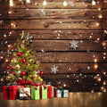 A decorated Christmas tree with red and gold ornaments and a star on top stands next to several wrapped gifts on a wooden floor; string lights and snowflake decorations adorn the ideasbackdrop Wood Photo Backdrop Snowflake Brown Wooden Wall Background Photography for Party Wedding Baby Photoshoot.