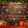 Wood wall backdrop decorated with string lights, snowflakes, and garlands. Snow falls gently in front of the wall. Wrapped Christmas presents and ornaments are arranged on a wooden surface below, creating a festive scene perfect for ideasbackdrop's Wood Photo Backdrop Snowflake Brown Wooden Wall Background Photography for Party Wedding Baby Photoshoot enthusiasts.