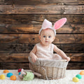 A baby wearing bunny ears sits in a wicker basket on a fluffy carpet, surrounded by colorful eggs and a toy, with an ideasbackdrop Brown Wood Backdrop Photographers for Birthday Baby Shower Background Photo Booth Video Shoot Studio Prop in the background.