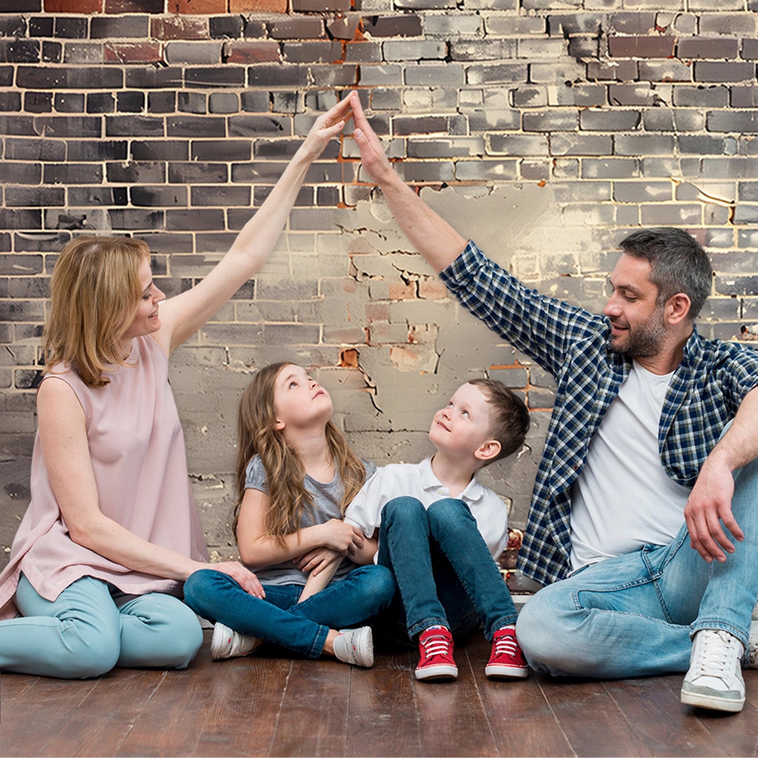 A family of four, seated on the floor against an ideasbackdrop Vintage Distressed Brick Wall Backdrop for Photography Portrait Background Studio Props, form an arch with their raised hands while the children look up, capturing a moment perfect for photo studio photography.