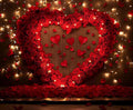 A large heart shape made of red roses is mounted on a wall, surrounded by string lights. Red rose petals and smaller heart decorations are scattered around, creating a Floral Wall Valentine's Day Flower Background - ideasbackdrop.