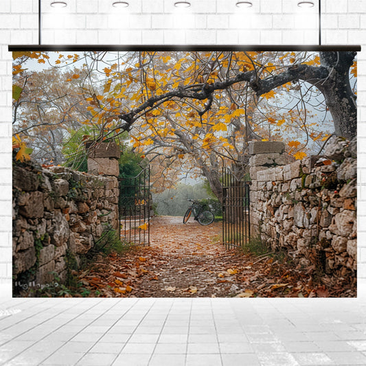 A stone pathway with autumn leaves leads through an open gate flanked by stone walls. A bicycle is parked on the path under trees with yellow and orange leaves, creating the perfect Gate Stone Wall Autumn Leaves Trees Fall Backdrop-ideasbackdrop for seasonal theme fall portraits by ideasbackdrop.