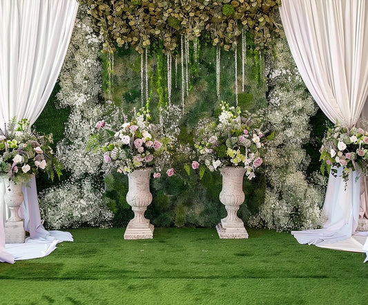 A decorated backdrop features white drapes, green foliage, and white flowers, creating a breathtaking floral atmosphere perfect for any special occasion. Three stone planters hold pink and white floral arrangements, while the floor is covered in green carpet, enhancing the Green Grassfield Wedding Flowers Backdrop -ideasbackdrop by ideasbackdrop.
