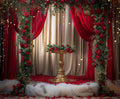 A gold pedestal is surrounded by red roses, red drapes, and fairy lights, set on a stage with red petals on the floor and complemented by a Red Floral Wedding Bridal Shower Backdrop -ideasbackdrop.