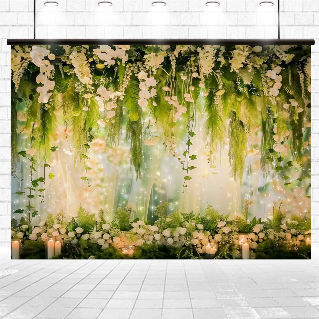 A Romantic Floral Weddings Photography Backdrop -ideasbackdrop from ideasbackdrop with green foliage and white flowers hangs from a white wall, creating an enchanting event decor. The setting is enhanced by hanging lights and candles, casting a soft, illuminated ambiance.