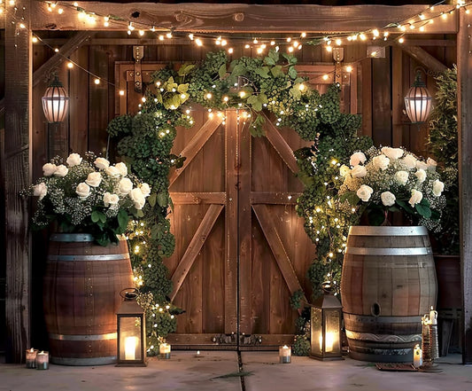 Outdoor rustic wedding set-up with wooden doors adorned with greenery and white flowers. String lights, lanterns, and candles create a warm ambiance. Two large floral arrangements on wine barrels showcase floral finesse against an HD vivid *Rustic Western Barn Door Floral Backdrop -ideasbackdrop* by ideasbackdrop.