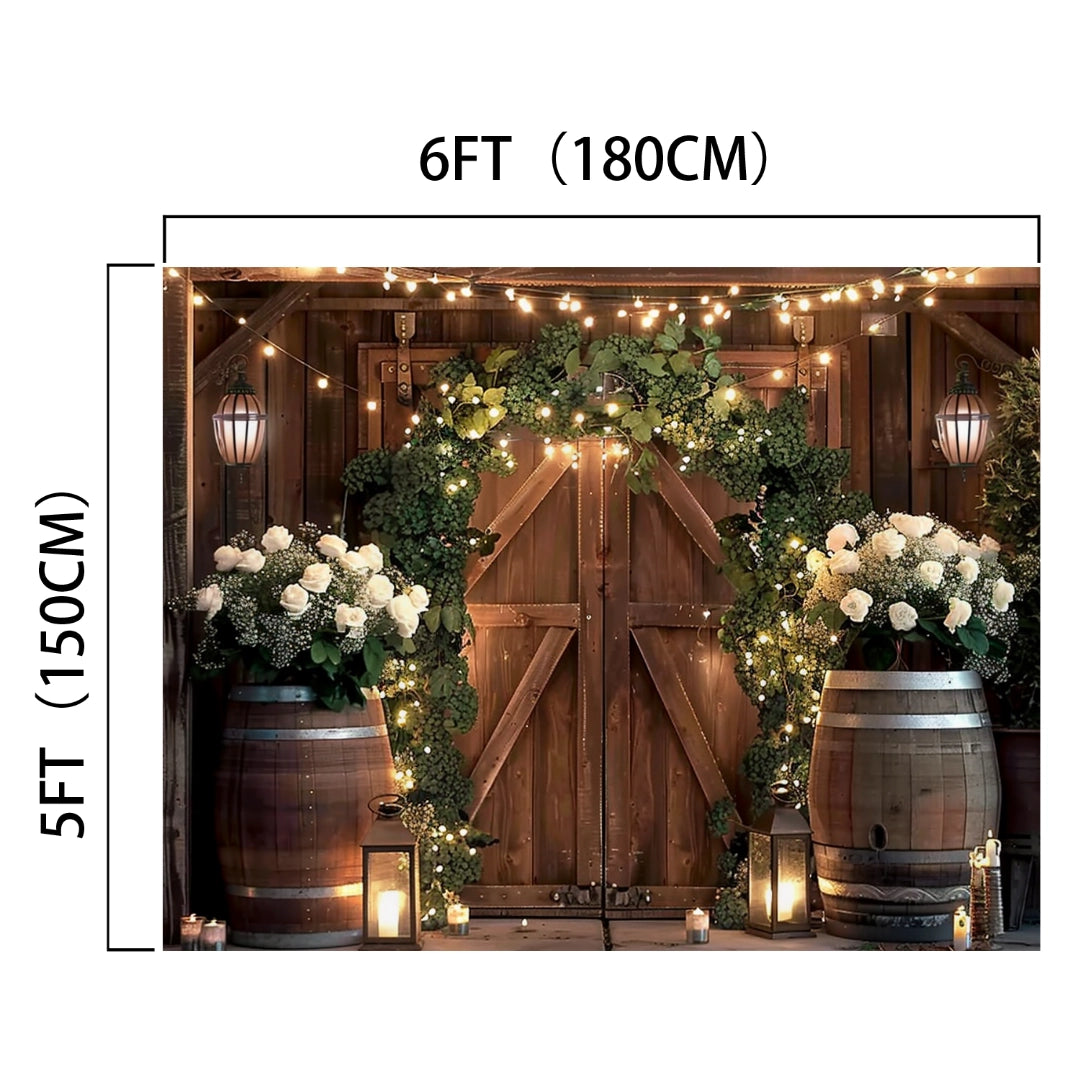 Backdrop of a rustic wooden door adorned with lifelike green foliage and white flowers, vibrant hues, barrels, and string lights, measuring 6 feet by 5 feet. Suitable for weddings or events, with lanterns and candles as decoration. Ideal for creating an ideasbackdrop Rustic Western Barn Door Floral Backdrop -ideasbackdrop.