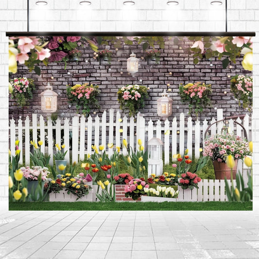 A white picket fence with flower pots, tulips, and lanterns adorns a green garden space against a Spring Garden Fence Floral Brick Wall Flower Backdrop-ideasbackdrop from ideasbackdrop, creating the perfect setting for photo shoots and events.