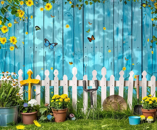 A blue wooden fence adorned with yellow flowers and butterflies, complemented by a white picket fence in front, creates an enchanting photo shoot backdrop. Various potted plants, garden tools, and a straw hat rest on the grass. The Spring Garden Wood Fence Flower Backdrop -ideasbackdrop by ideasbackdrop ensures every detail is captured in high-definition quality.