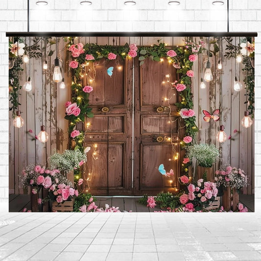 A rustic wooden door adorned with ultra-realistic floral designs of pink roses, greenery, string lights, and butterflies. The setting includes floral arrangements on crates and Spring Photography Backdrop Wooden Barn Door -ideasbackdrop with hanging lights above—perfect for event planners seeking a magical touch.