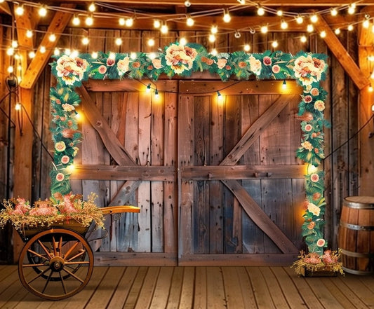 A picturesque countryside barn door adorned with floral garlands and string lights exudes rustic elegance decor. A wooden cart brimming with flowers and a barrel on the right side complete this charming scene, perfect for a Western Cowboy Floral Wood Barn Barn Backdrop by ideasbackdrop.