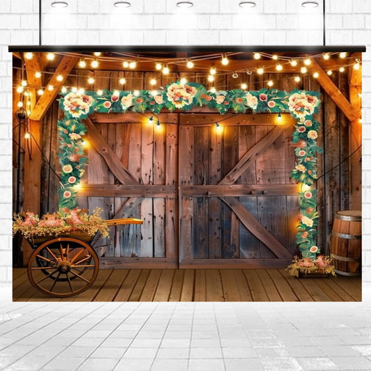 A rustic wooden barn door adorned with string lights and floral garlands, set against a patio area with a flower-filled wooden cart and barrels, capturing rustic elegance reminiscent of a picturesque countryside barn. This Western Cowboy Floral Wood Barn Backdrop-ideasbackdrop by ideasbackdrop adds charm to any setting.