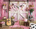 A rustic setup featuring a weathered barn door, hay bales, a guitar, cowboy boots, pink decor, flowers, a wagon wheel, and a milk bucket. Triangular pennants hang above on an HD vivid ideasbackdrop Western Cowgirl Wild West Rustic Barn Backdrop that exudes rustic beauty with its pink wood background.