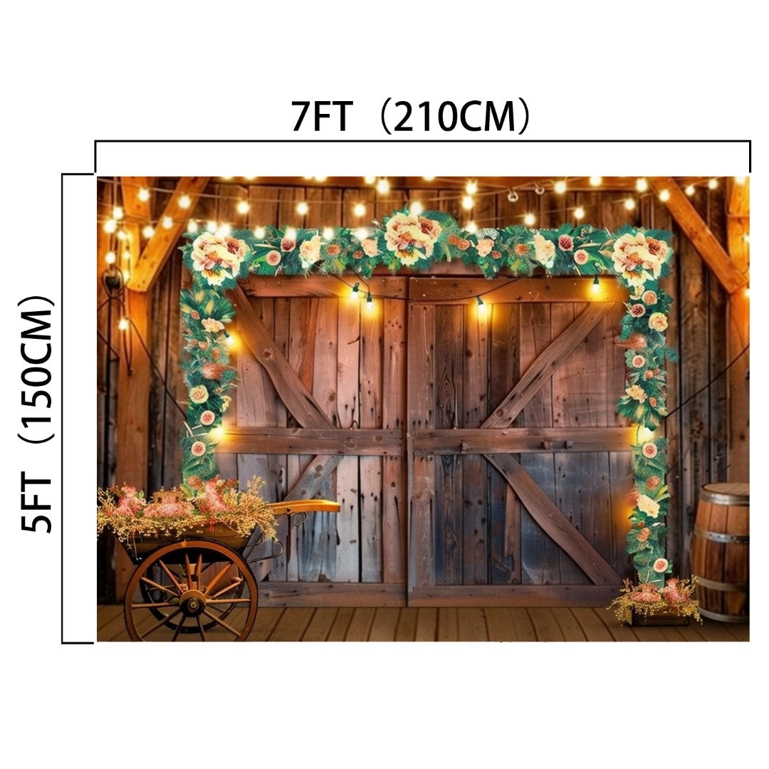 Wooden barn doors adorned with string lights and floral garlands, exuding rustic elegance. Dimensions are 7 feet (210 cm) wide and 5 feet (150 cm) tall. A small cart with flowers sits on the left, and a barrel is on the right, creating an ideasbackdrop Western Cowboy Floral Wood Barn Barn Backdrop-ideasbackdrop perfect for a countryside barn setting.