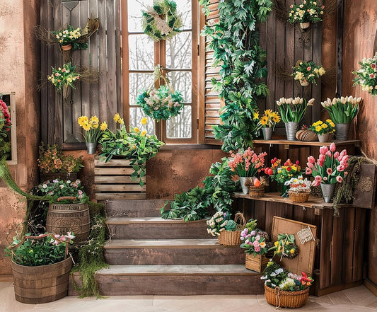 A rustic indoor setting with wooden stairs, walls, and various potted plants and flowers, including tulips, daffodils, and ivy. Complemented by a Wooden Window Garden Grass Floral Backdrop-ideasbackdrop by ideasbackdrop and a window with a snowy view in the background, it's perfect for events and photography.